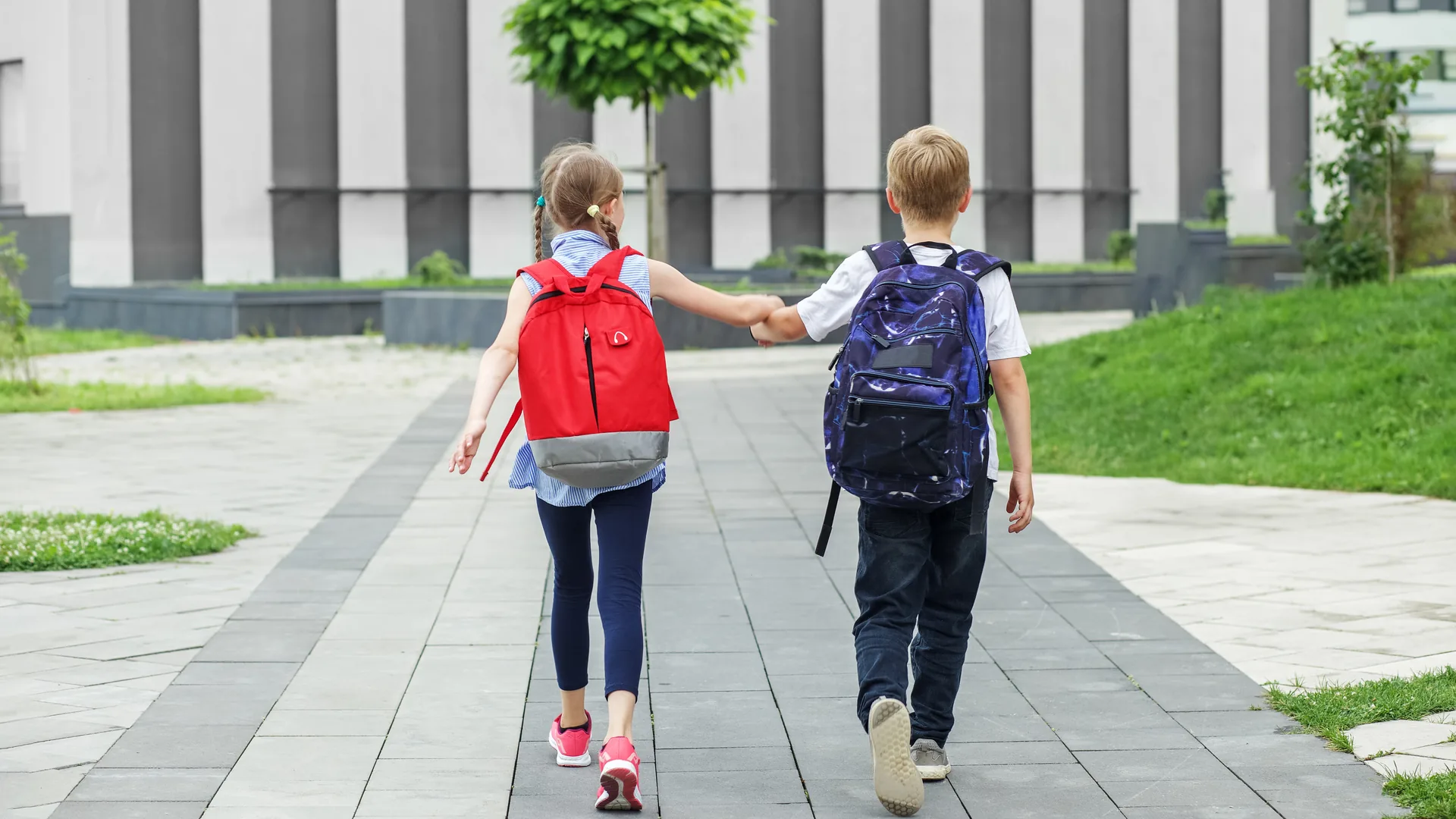 To skolebarn med sekker på vei til skolen hånd i hånd, sett bakfra