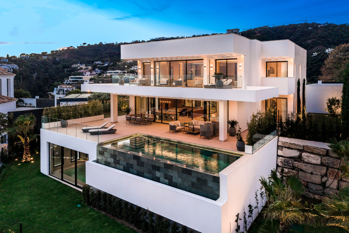 Villa Alfa – El Madroñal - moderne hvid facade med infinity-pool, terrasser og bakkeudsigt i skumringen