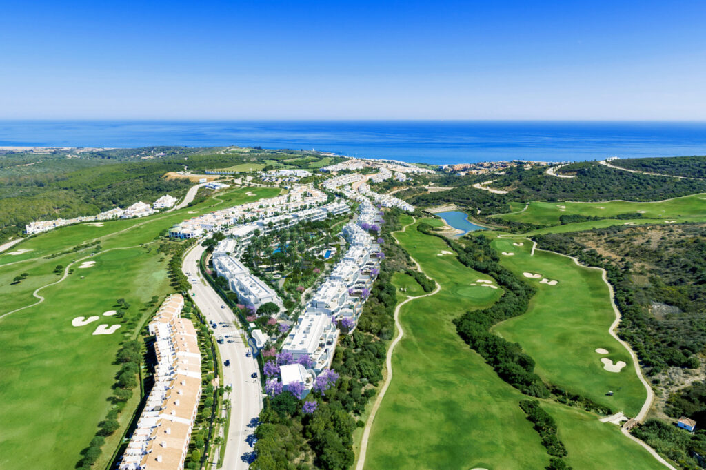 Serenity Frontline Golf Homes and Beach Club – PHASE IV – La Alcaidesa - aerial view of white residential complex flanked by lush golf fairways with Mediterranean Sea backdrop