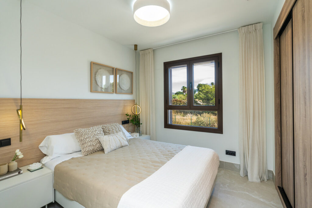 The Meadows – FASE 1 – La Cala Golf - elegantly styled bedroom with wood headboard, pendant lights, and garden views through dark-framed window