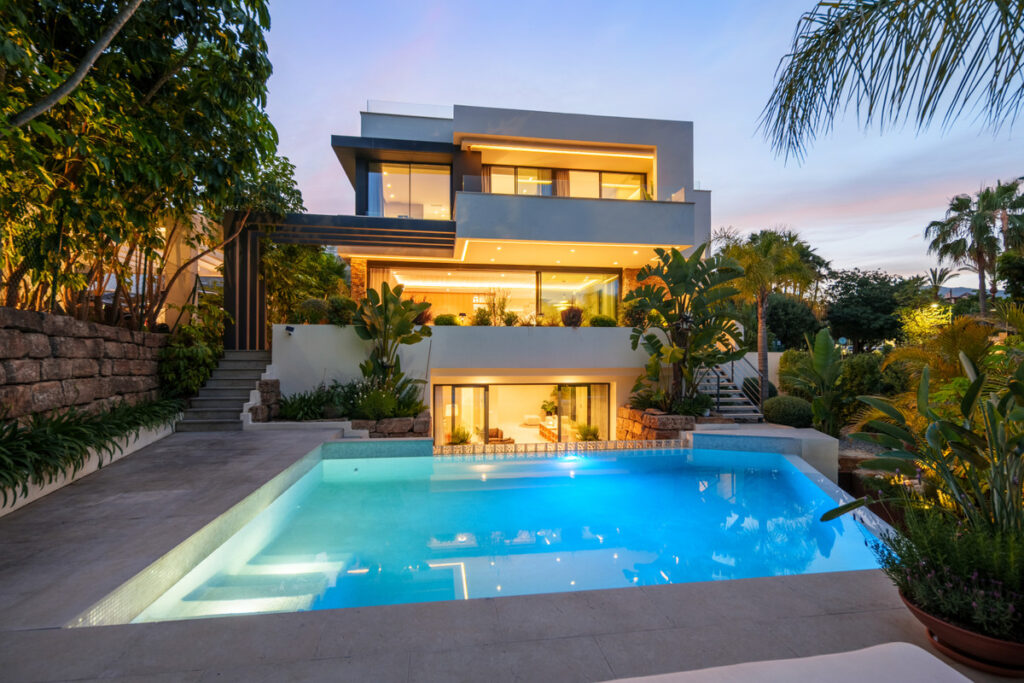 Belfry 1 – Estepona - moderne dreistöckige Luxusvilla mit beleuchtetem Pool in der Abenddämmerung, umgeben von tropischen Gärten